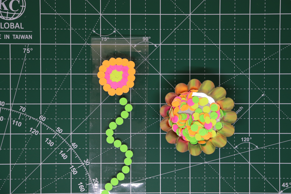 A container of colorful paper dots, by packing tape with a green paper dot stem and yellow, pink, and orange paper dots in concentric circes making the beginning of a flower