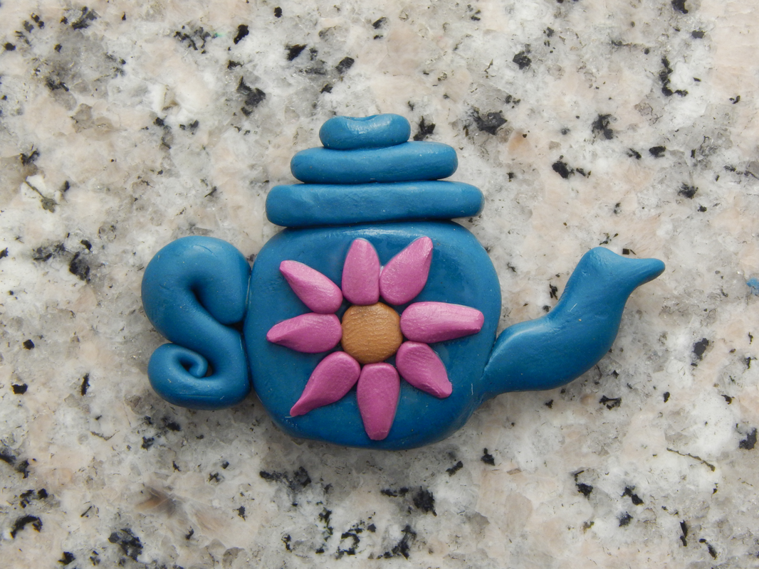 A blue teapot bead on a grey speckled background with a pink flower with a brown center added to it. 