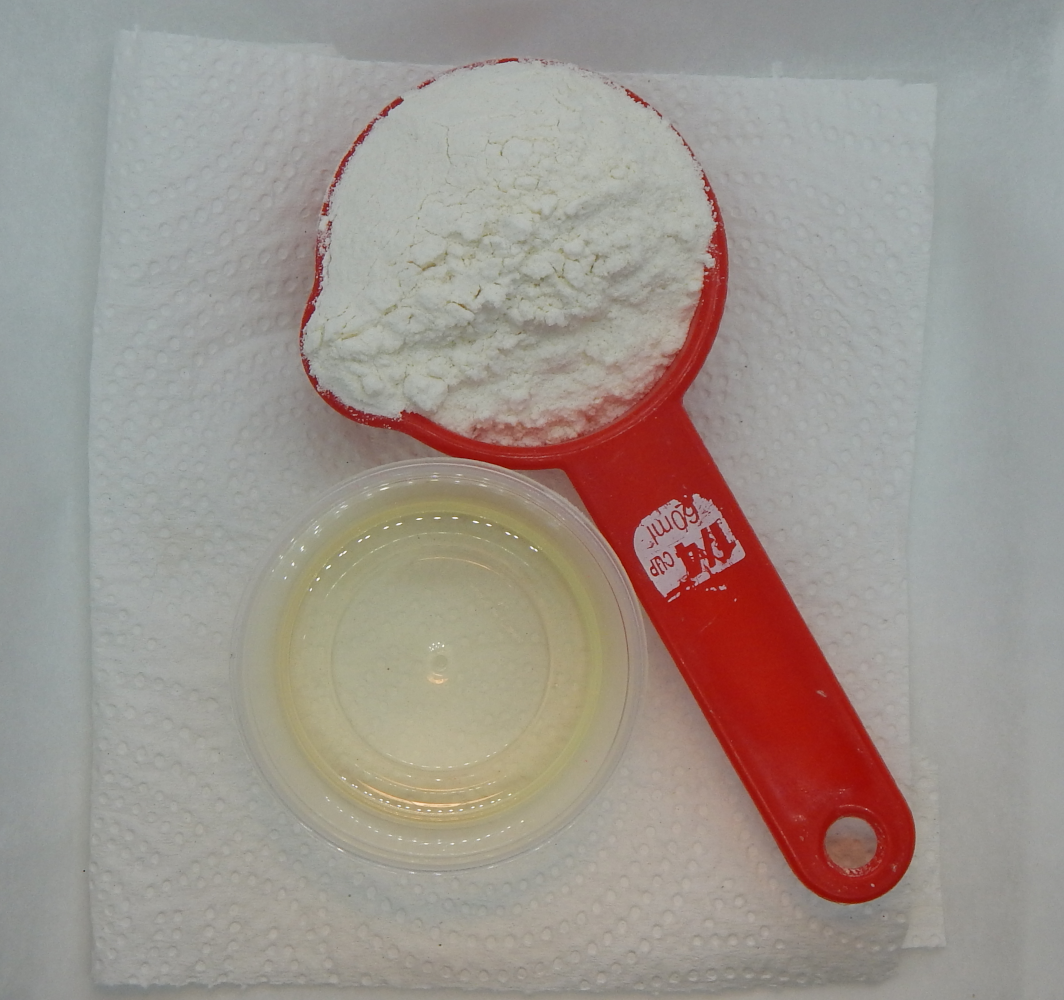 a red measuring cup holding a mound of flour, a small plastic cup holding cooking oil.