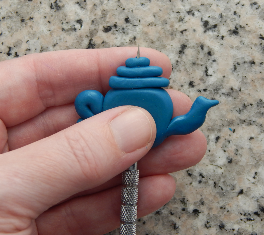 A hand gently holds a teapot bead while a needle tool is inserted from bottom to top