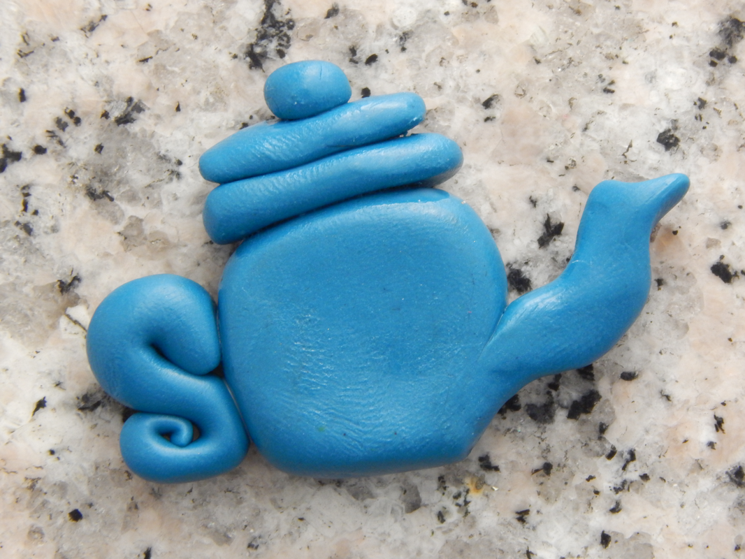 A blue polymer clay teapot (miniature/bead sized) on grey speckled background