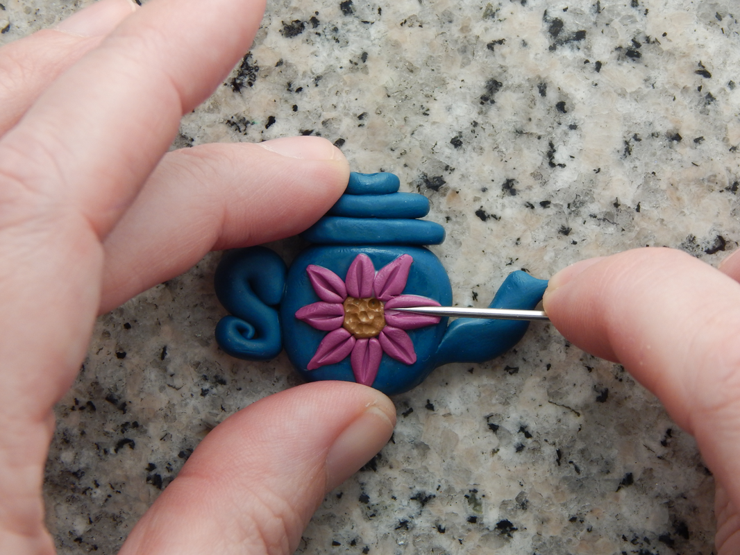 One hand holds the teapot bead while the other presses a needle tool in the middle of one of the pink flower petals. The rest of the flower petals have also been textured in this way.